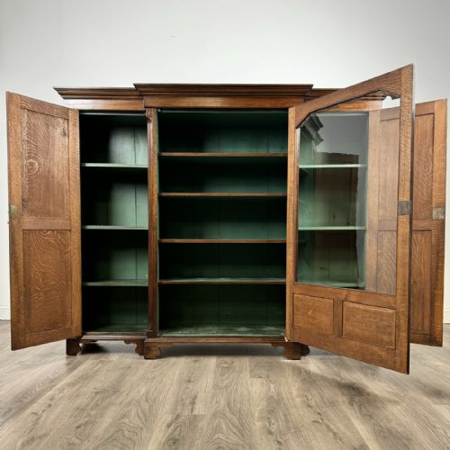 fig 1. A large dark oak display cabinet featuring a central glazed door with shelves, flanked by two solid panelled doors adorned with decorative brass strap hinges.
