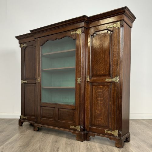 fig 2. A large dark oak display cabinet featuring a central glazed door with shelves, flanked by two solid panelled doors adorned with decorative brass strap hinges.