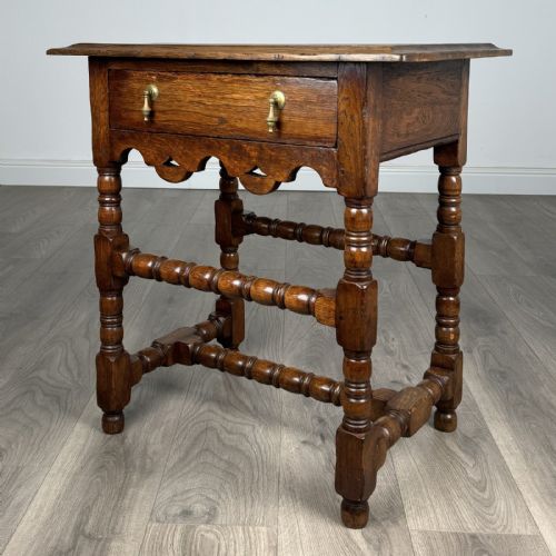 fig 1. An antique dark oak side table featuring a single drawer with brass drop handles, a scalloped apron, and bobbin-turned legs connected by matching stretchers.