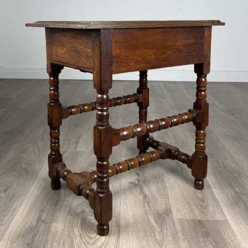 fig 3. An antique dark oak side table featuring a single drawer with brass drop handles, a scalloped apron, and bobbin-turned legs connected by matching stretchers.