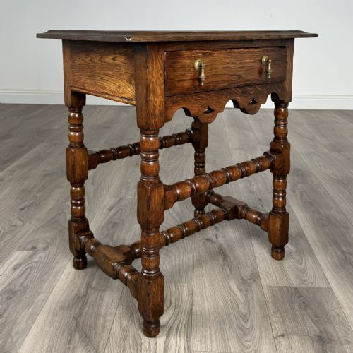 fig 7. An antique dark oak side table featuring a single drawer with brass drop handles, a scalloped apron, and bobbin-turned legs connected by matching stretchers.