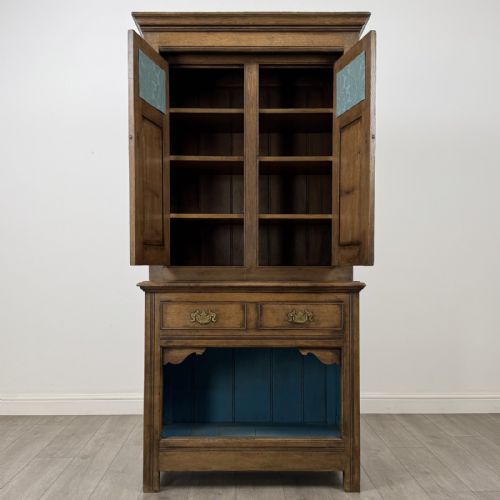 fig 1. A tall antique brown oak cupboard with carved spindle panels on the upper cabinet doors, brass butterfly hinges, two drawers, and a lower open alcove with a blue painted interior.
