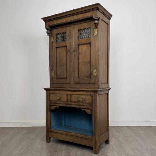 fig 2. A tall antique brown oak cupboard with carved spindle panels on the upper cabinet doors, brass butterfly hinges, two drawers, and a lower open alcove with a blue painted interior.