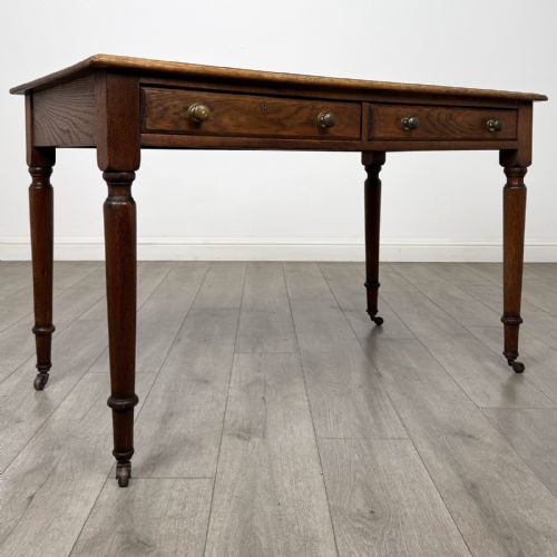 fig 7. An antique golden oak writing table featuring two frieze drawers with turned knobs and supported by turned legs on brass castors.