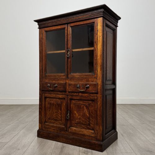 fig 2. A dark oak Victorian display cabinet featuring a dentil cornice, glazed upper doors, two drawers with brass handles, and panelled lower cupboard doors.