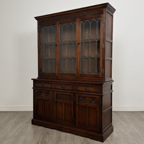 fig 2. A dark oak display cabinet featuring three leaded glass doors, three drawers, and three carved panelled cupboards with brass handles.