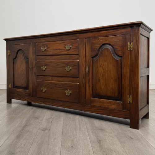 fig 2. A dark oak sideboard featuring three central drawers flanked by two arched panel cupboard doors with brass handles.