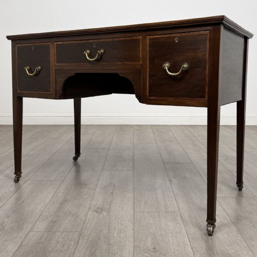 fig 1. An Edwardian mahogany writing table featuring three inlaid drawers, brass drop handles, square tapered legs, and brass castors.