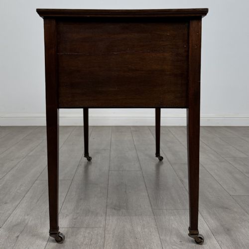 fig 6. An Edwardian mahogany writing table featuring three inlaid drawers, brass drop handles, square tapered legs, and brass castors.