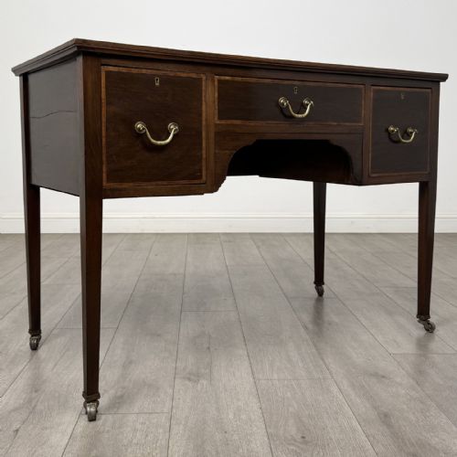 fig 7. An Edwardian mahogany writing table featuring three inlaid drawers, brass drop handles, square tapered legs, and brass castors.