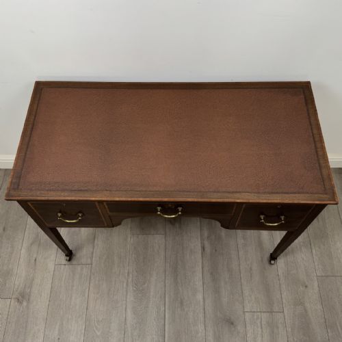 fig 8. An Edwardian mahogany writing table featuring three inlaid drawers, brass drop handles, square tapered legs, and brass castors.