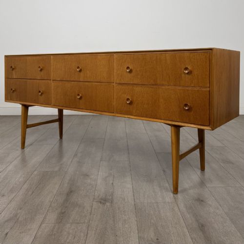fig 1. A mid-century oak sideboard featuring six wide drawers with circular wooden pull knobs, supported by tapered legs and side stretchers.
