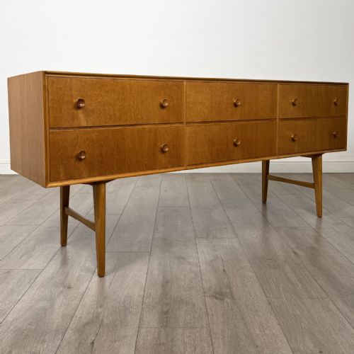 fig 7. A mid-century oak sideboard featuring six wide drawers with circular wooden pull knobs, supported by tapered legs and side stretchers.