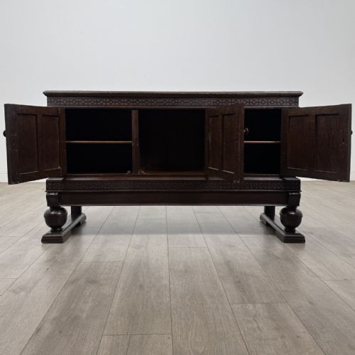 fig 1. A dark oak Edwardian low cupboard featuring three panelled doors, carved frieze borders, and turned bulbous legs on sledge feet.