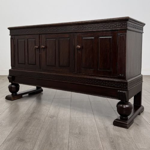 fig 2. A dark oak Edwardian low cupboard featuring three panelled doors, carved frieze borders, and turned bulbous legs on sledge feet.