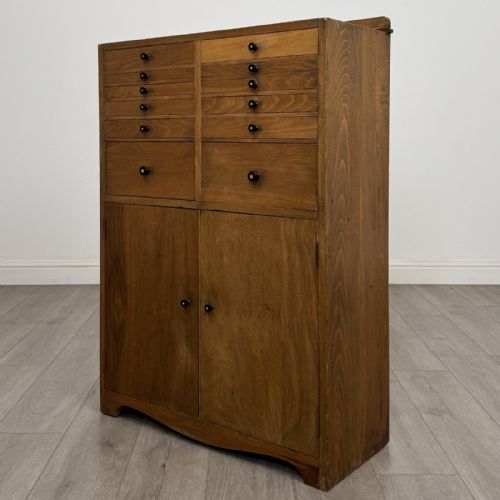 fig 2. An antique oak storage cabinet featuring multiple shallow drawers arranged above a double-door lower cupboard with small turned dark wooden knobs.