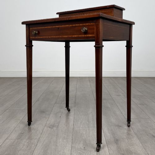fig 1. An Edwardian mahogany writing table featuring boxwood inlay, a single drawer with brass knobs, a raised rear stationery shelf, and square tapered legs on castors.