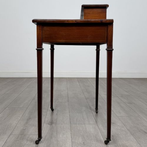 fig 2. An Edwardian mahogany writing table featuring boxwood inlay, a single drawer with brass knobs, a raised rear stationery shelf, and square tapered legs on castors.