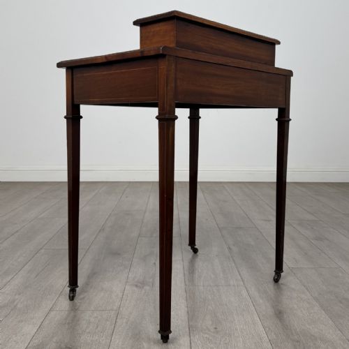 fig 3. An Edwardian mahogany writing table featuring boxwood inlay, a single drawer with brass knobs, a raised rear stationery shelf, and square tapered legs on castors.