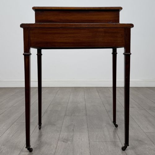 fig 4. An Edwardian mahogany writing table featuring boxwood inlay, a single drawer with brass knobs, a raised rear stationery shelf, and square tapered legs on castors.