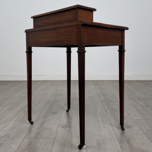 fig 5. An Edwardian mahogany writing table featuring boxwood inlay, a single drawer with brass knobs, a raised rear stationery shelf, and square tapered legs on castors.