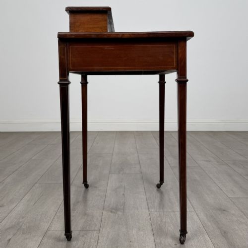 fig 6. An Edwardian mahogany writing table featuring boxwood inlay, a single drawer with brass knobs, a raised rear stationery shelf, and square tapered legs on castors.