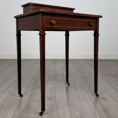 fig 7. An Edwardian mahogany writing table featuring boxwood inlay, a single drawer with brass knobs, a raised rear stationery shelf, and square tapered legs on castors.