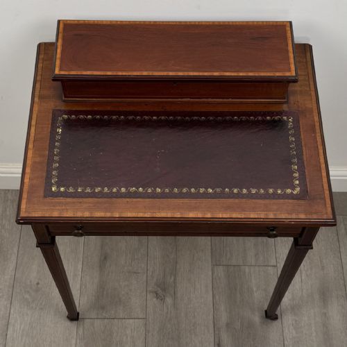 fig 8. An Edwardian mahogany writing table featuring boxwood inlay, a single drawer with brass knobs, a raised rear stationery shelf, and square tapered legs on castors.