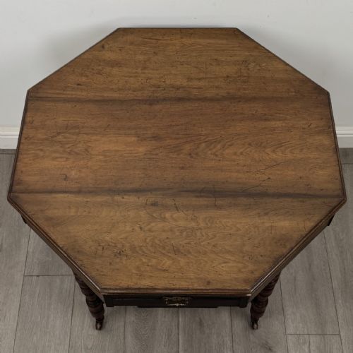 fig 6. An antique dark mahogany octagonal centre table featuring a drawer with a brass drop handle, ring-turned legs on brass castors, and an octagonal lower shelf with a spindle gallery.