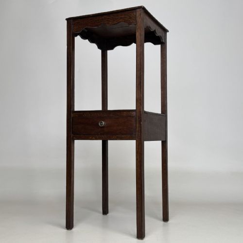fig 1. A tall dark oak Edwardian side table featuring a top surface with a scalloped apron shelf beneath, a single drawer with a central brass knob, and four slender square legs.