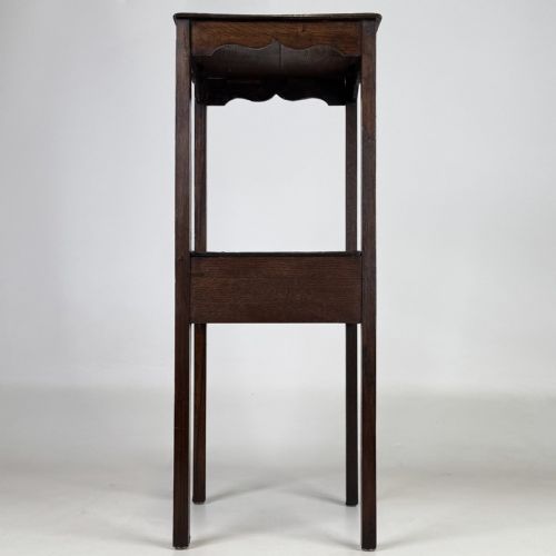 fig 2. A tall dark oak Edwardian side table featuring a top surface with a scalloped apron shelf beneath, a single drawer with a central brass knob, and four slender square legs.