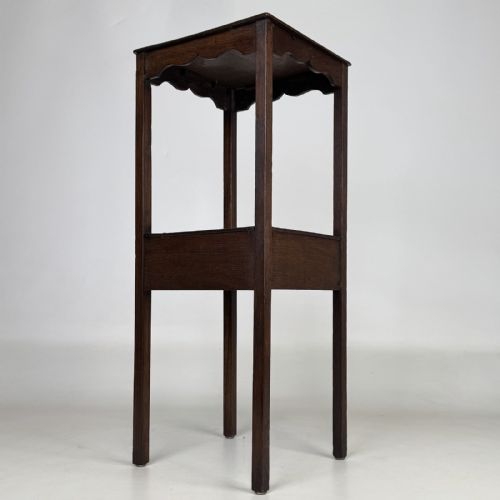 fig 3. A tall dark oak Edwardian side table featuring a top surface with a scalloped apron shelf beneath, a single drawer with a central brass knob, and four slender square legs.