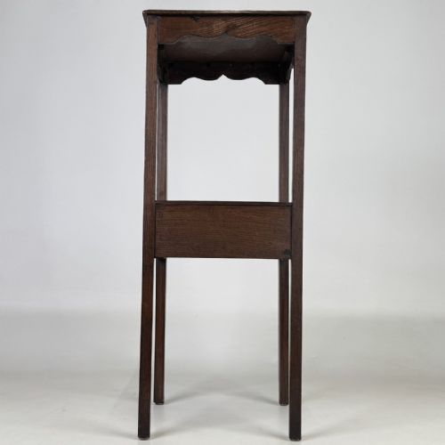 fig 4. A tall dark oak Edwardian side table featuring a top surface with a scalloped apron shelf beneath, a single drawer with a central brass knob, and four slender square legs.