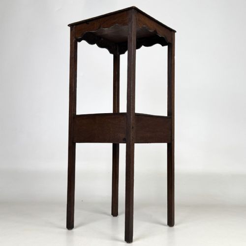 fig 5. A tall dark oak Edwardian side table featuring a top surface with a scalloped apron shelf beneath, a single drawer with a central brass knob, and four slender square legs.