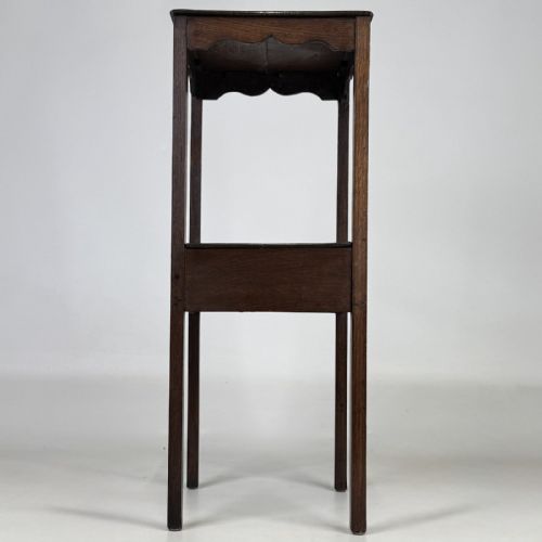 fig 6. A tall dark oak Edwardian side table featuring a top surface with a scalloped apron shelf beneath, a single drawer with a central brass knob, and four slender square legs.