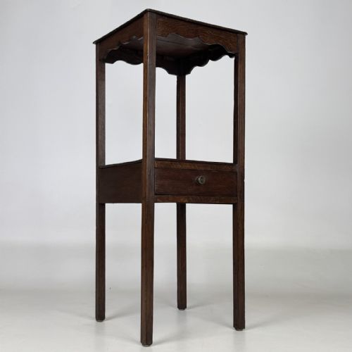 fig 7. A tall dark oak Edwardian side table featuring a top surface with a scalloped apron shelf beneath, a single drawer with a central brass knob, and four slender square legs.