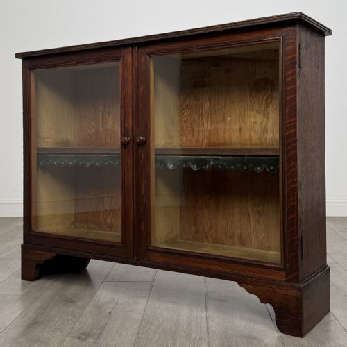 fig 3. A low Victorian dark oak display cabinet featuring two glazed doors, turned wooden knobs, bracket feet, and a single interior shelf trimmed with scalloped leather.