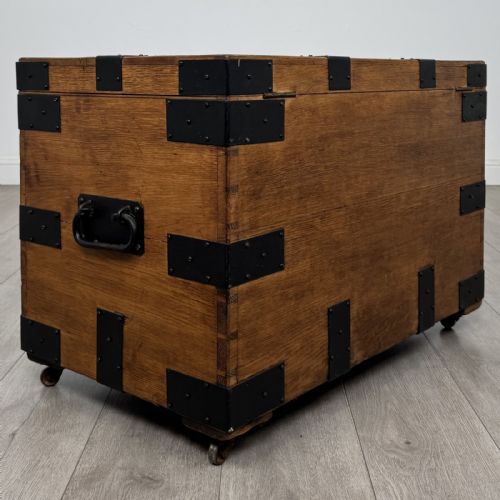 fig 3. A rectangular warm brown oak storage chest featuring black metal strap reinforcement, a keyhole, and corner brackets on small castors.