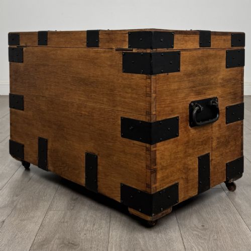 fig 5. A rectangular warm brown oak storage chest featuring black metal strap reinforcement, a keyhole, and corner brackets on small castors.