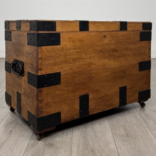 fig 7. A rectangular warm brown oak storage chest featuring black metal strap reinforcement, a keyhole, and corner brackets on small castors.