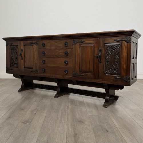 fig 2. A large dark carved oak sideboard featuring panelled cupboard doors with iron strap hinges, central drawers, and a carved low stretcher base.