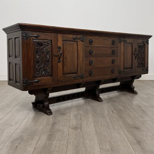fig 8. A large dark carved oak sideboard featuring panelled cupboard doors with iron strap hinges, central drawers, and a carved low stretcher base.