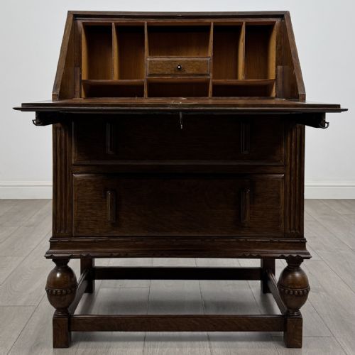 fig 1. An antique dark oak fall-front writing bureau featuring a carved oval motif on the sloping lid, two lower drawers with wooden handles, and bulbous turned front legs on a stretcher base.