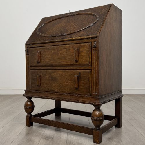 fig 2. An antique dark oak fall-front writing bureau featuring a carved oval motif on the sloping lid, two lower drawers with wooden handles, and bulbous turned front legs on a stretcher base.