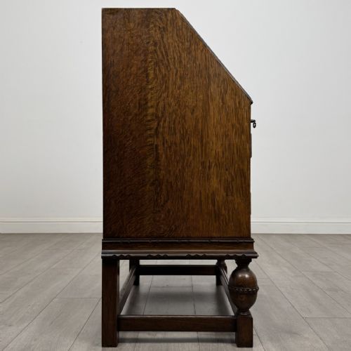 fig 7. An antique dark oak fall-front writing bureau featuring a carved oval motif on the sloping lid, two lower drawers with wooden handles, and bulbous turned front legs on a stretcher base.