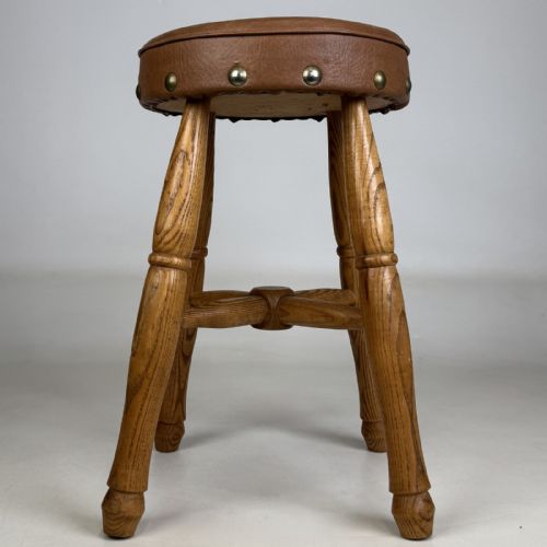 fig 5. A circular antique oak stool featuring a padded brown leather seat secured with brass studs, supported by four turned legs and cross stretchers.