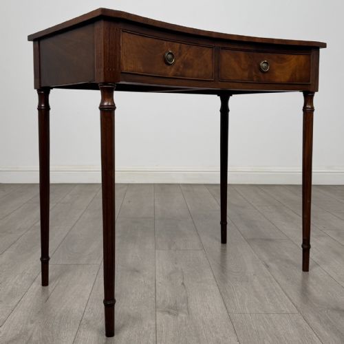 fig 7. An antique mahogany writing table with a concave bowfront shape, two drawers, brass ring handles, and four reeded legs.