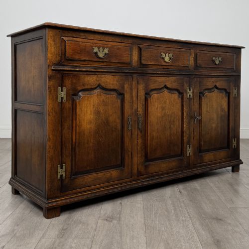 fig 7. A dark oak sideboard featuring three top drawers with brass handles above three fielded panel cabinet doors with exposed brass H-hinges.
