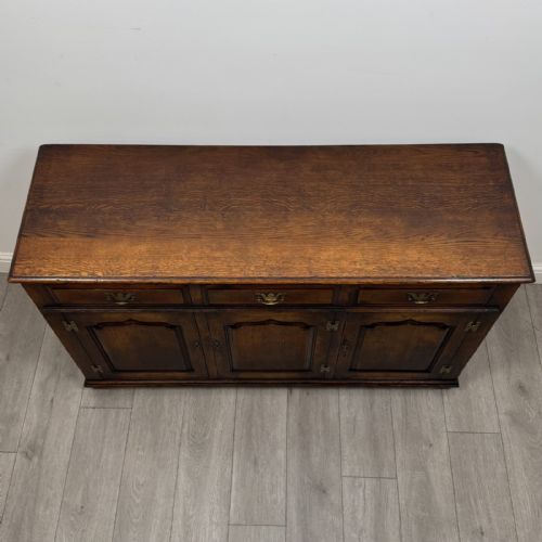 fig 8. A dark oak sideboard featuring three top drawers with brass handles above three fielded panel cabinet doors with exposed brass H-hinges.