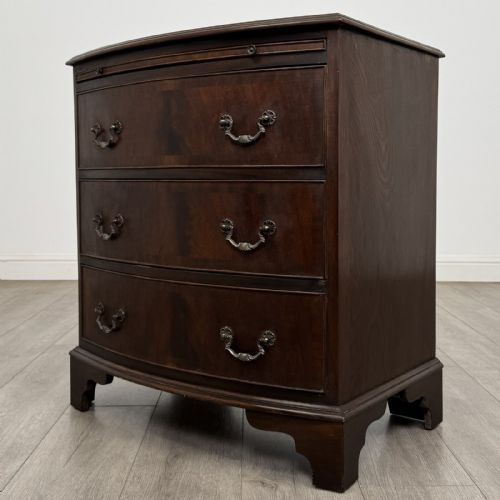 fig 1. A small dark mahogany bow-front chest of drawers featuring three drawers, a top pull-out slide, and brass swan-neck handles set on bracket feet.